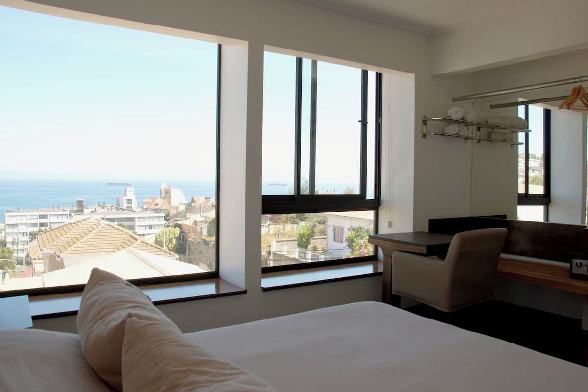 Camera d'albergo con un letto, una scrivania e una sedia. Ampie finestre offrono vista sulla città e sul mare.