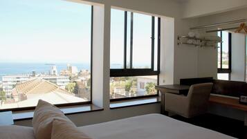 Ein Hotelzimmer mit Bett, Schreibtisch und Stuhl. Große Fenster bieten Blick auf die Stadt und das Meer.