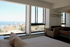 Habitación de hotel con cama, escritorio y silla. Amplias ventanas ofrecen vistas a la ciudad y al mar.