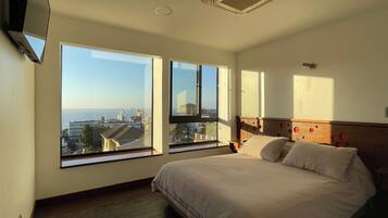 Un dormitorio con una cama grande, dos ventanas con vistas a la ciudad, un respiradero en el techo y un televisor montado en la pared.