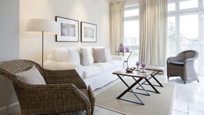En moderne stue med en hvid sofa, to fletstole, et sofabord og en gulvlampe. Der er to indrammede kunstværker på væggen og et vindue med gardiner.