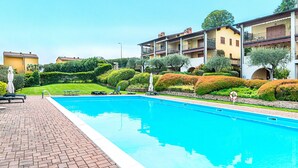 Une piscine entourée d’une terrasse en briques, bordée de haies et d’arbres bien entretenus, et située à côté d’un immeuble à plusieurs étages doté de balcons.