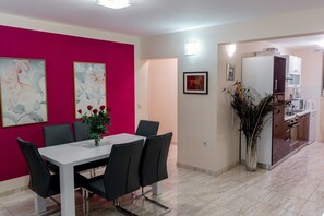 A modern dining area with a white table and black chairs, a vibrant pink wall with floral artwork, and a kitchen area with a refrigerator and coffee maker.