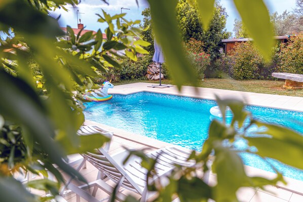 A swimming pool with clear blue water, surrounded by greenery and a wooden deck. - Private pool, 500 meters from the ocean, and two apartments for two families (Peroj)