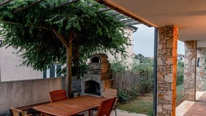 Une terrasse extérieure dotée d’une cheminée en pierre, d’une table et de chaises en bois, ainsi que d’une pergola couverte de plantes grimpantes.