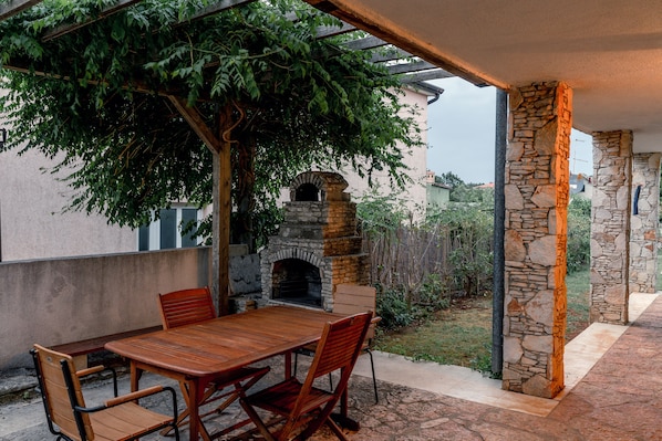 An outdoor patio with a stone fireplace, wooden table and chairs, and a pergola with climbing plants.