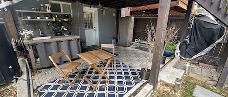 Un espacio de patio cubierto con mesa y sillas de madera, un tapete estampado y un pequeño jardín con plantas en macetas.