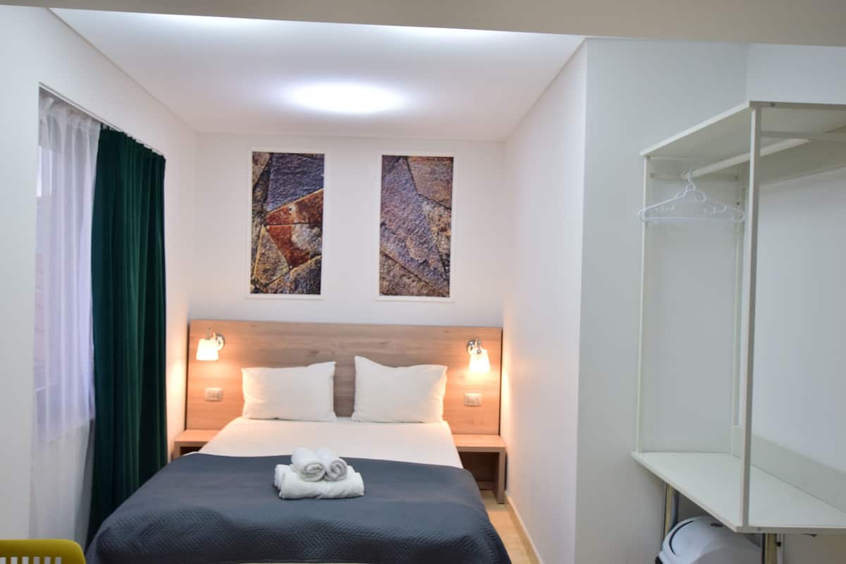 Ein ordentlich eingerichtetes Hotelzimmer mit einem großen Bett, zwei an der Wand befestigten Kunstwerken, einem Nachttisch mit einer Lampe und einem Kleiderschrank mit Kleiderbügel.