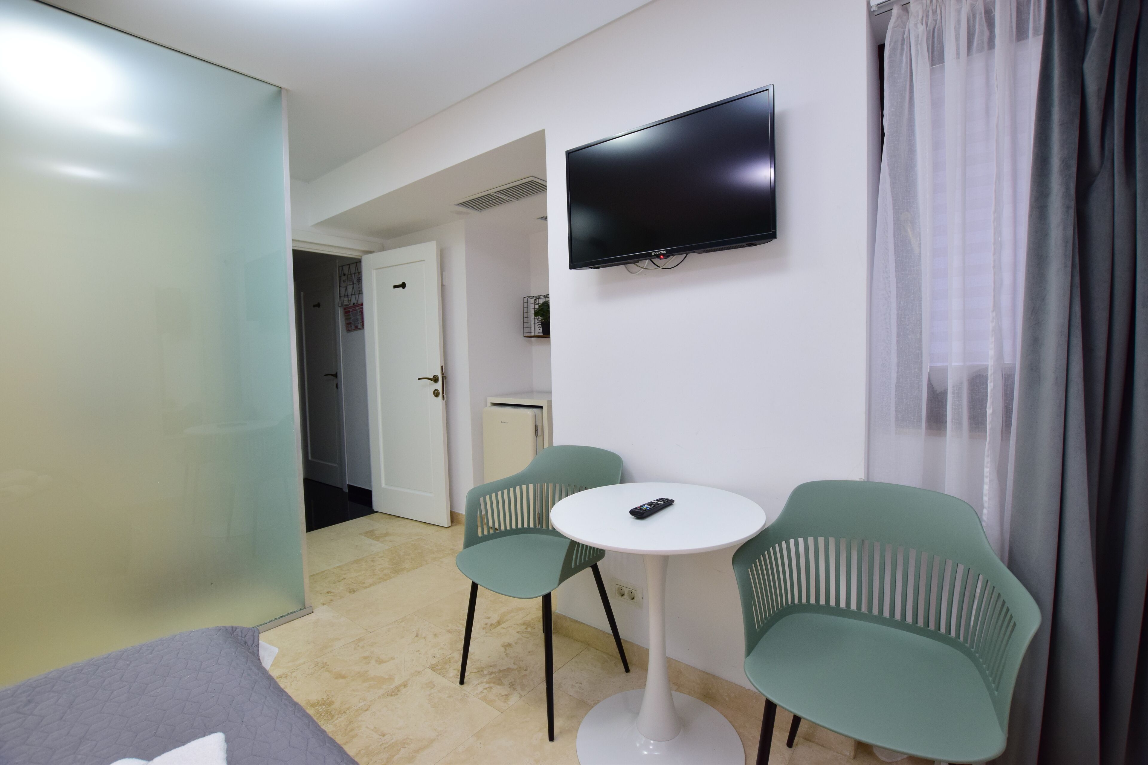 A hotel room with a bed, a small table with two chairs, a TV mounted on the wall, and a door leading to a bathroom.