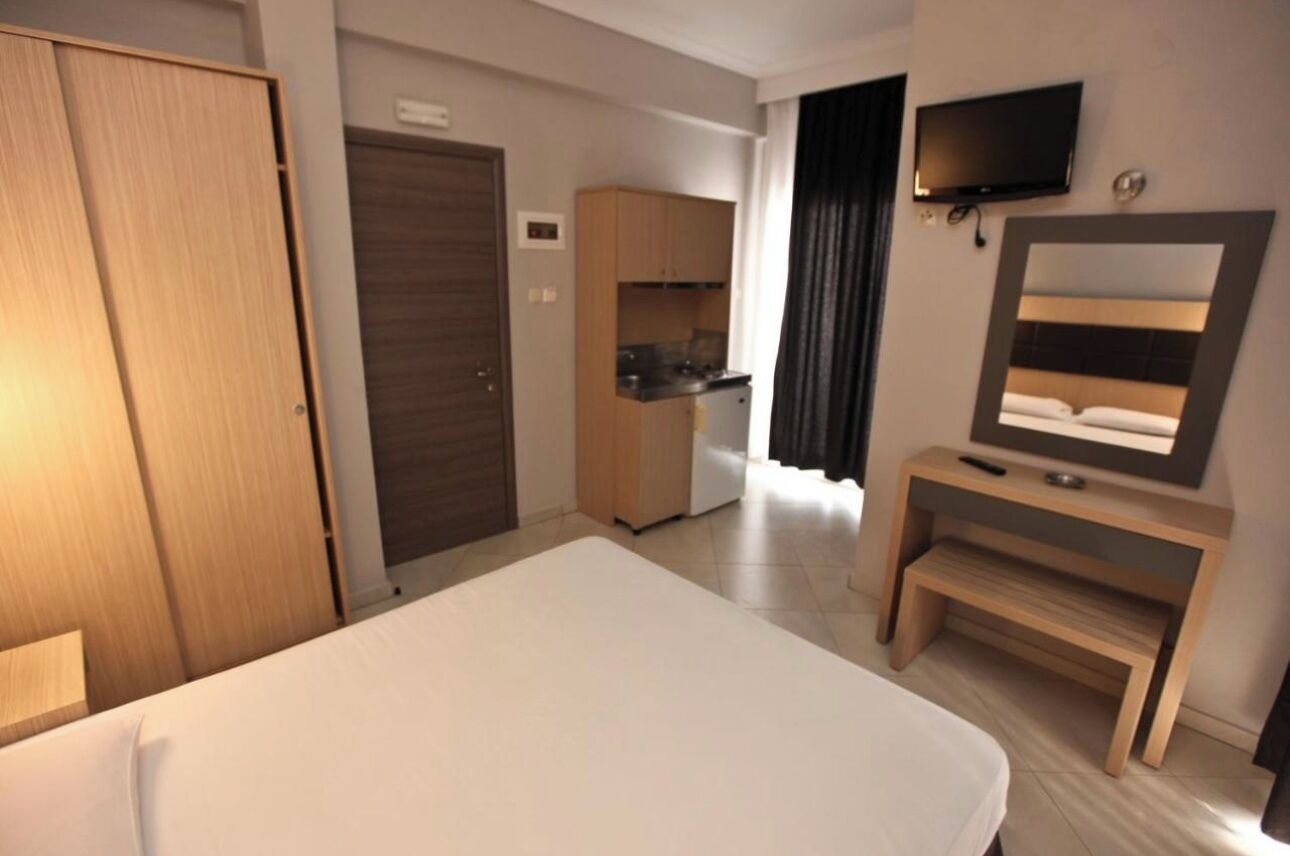 A hotel room with a bed, a TV mounted on the wall, a small desk with a mirror, and a kitchenette in the background.