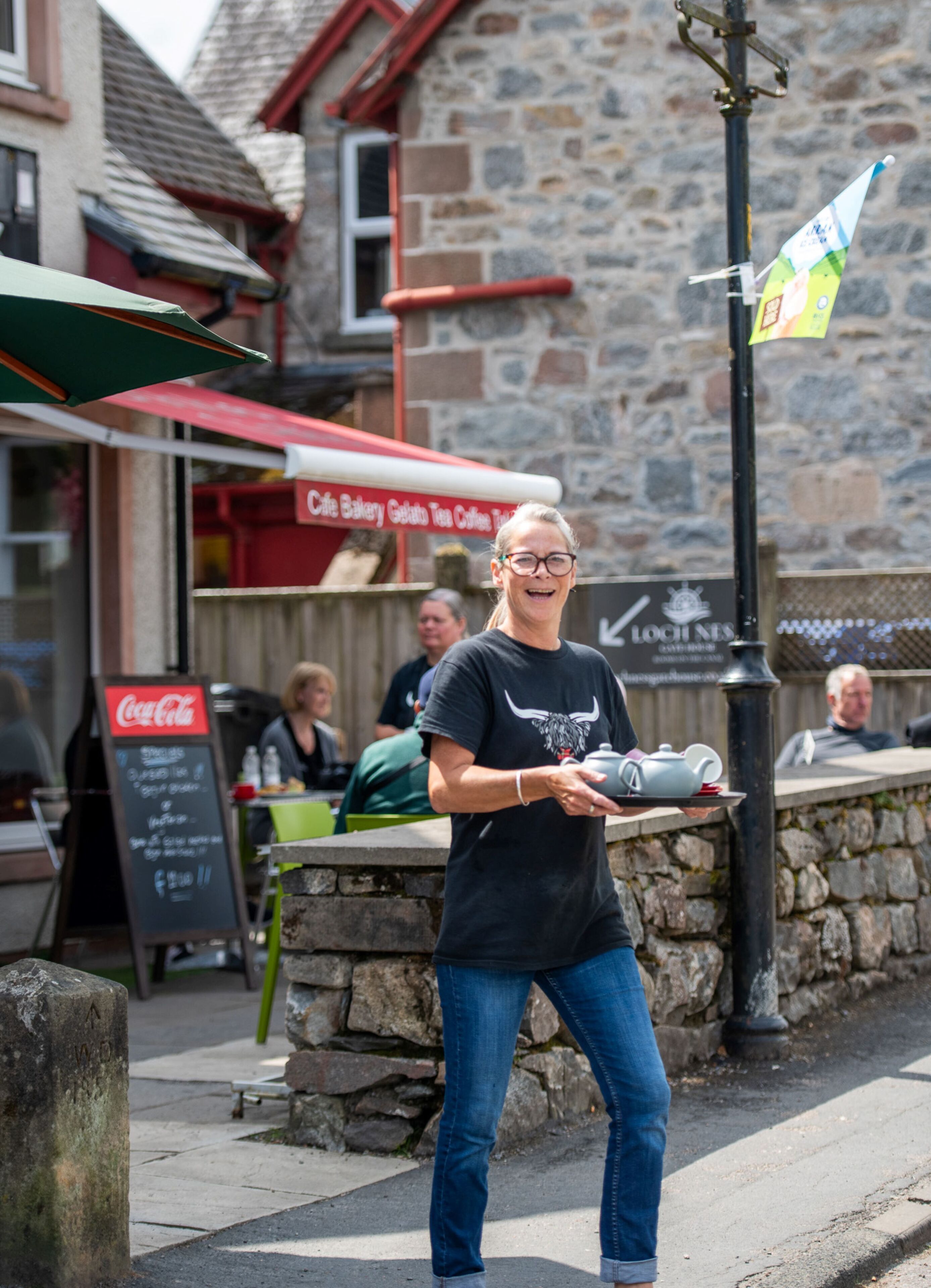 A person carrying a tray with a teapot and cup, standing in front of a stone building with a sign that reads "Lochines."
