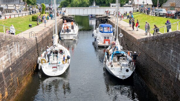 A canal with boats docked, a bridge in the background, and people walking along the banks. - Loch Ness Gate House (Fort Augustus)