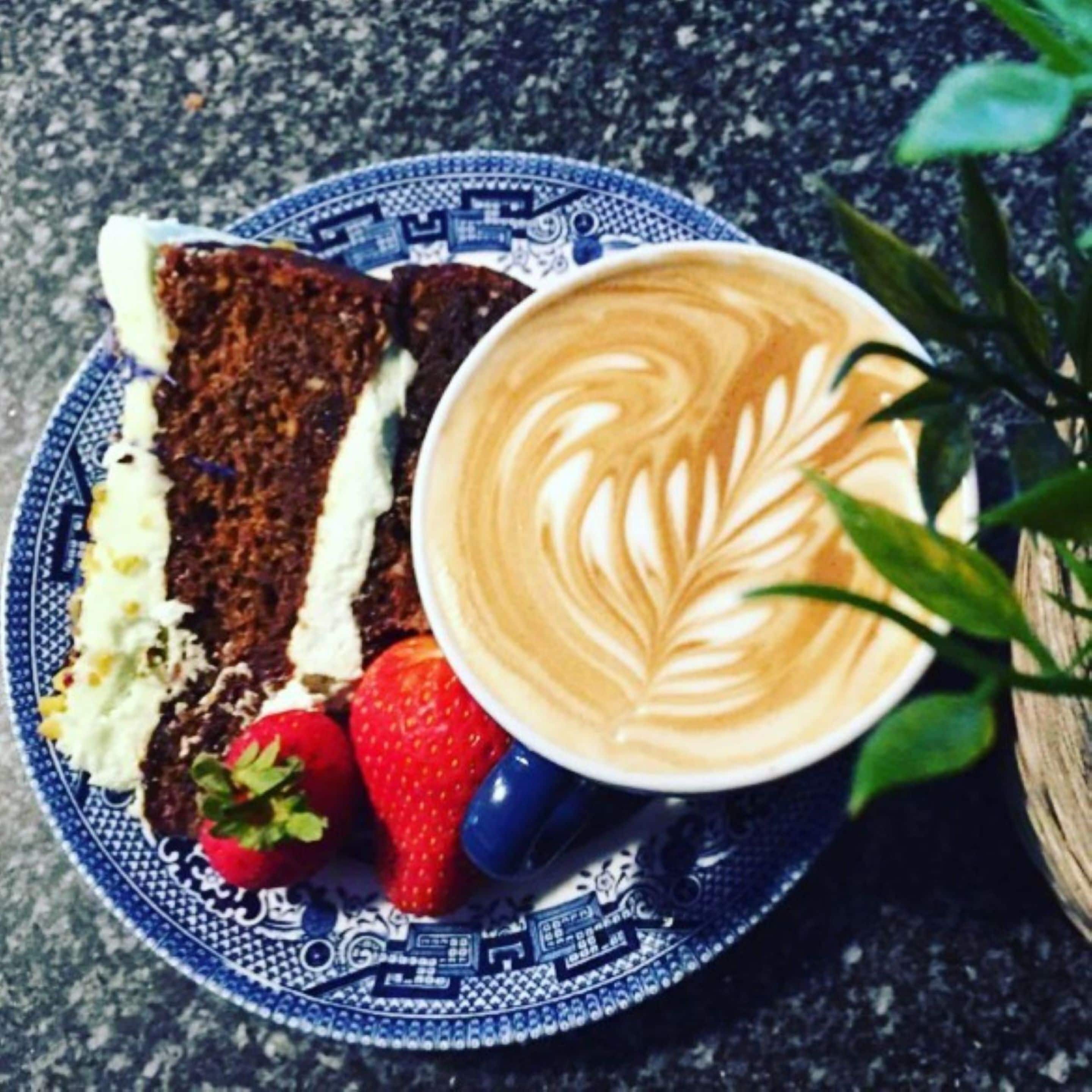 A plate with a slice of dark bread topped with white spread and a strawberry, accompanied by a cup of coffee with latte art.