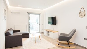 A modern living room with a grey sofa, a white coffee table, a wooden rocking chair, a TV mounted on the wall, and a large window. - Cozy and spacious apartment in Zografou (Zografos)