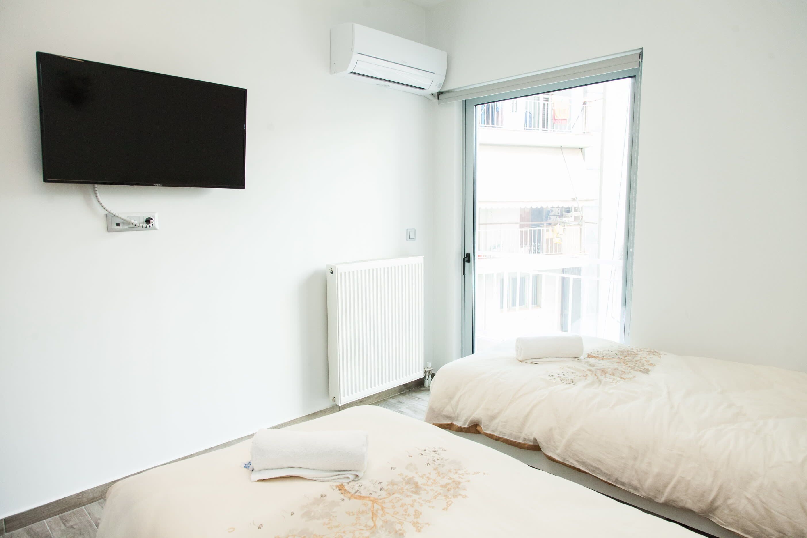 A bedroom with a bed, a TV, an air conditioner, a radiator, and a window with a view of buildings.