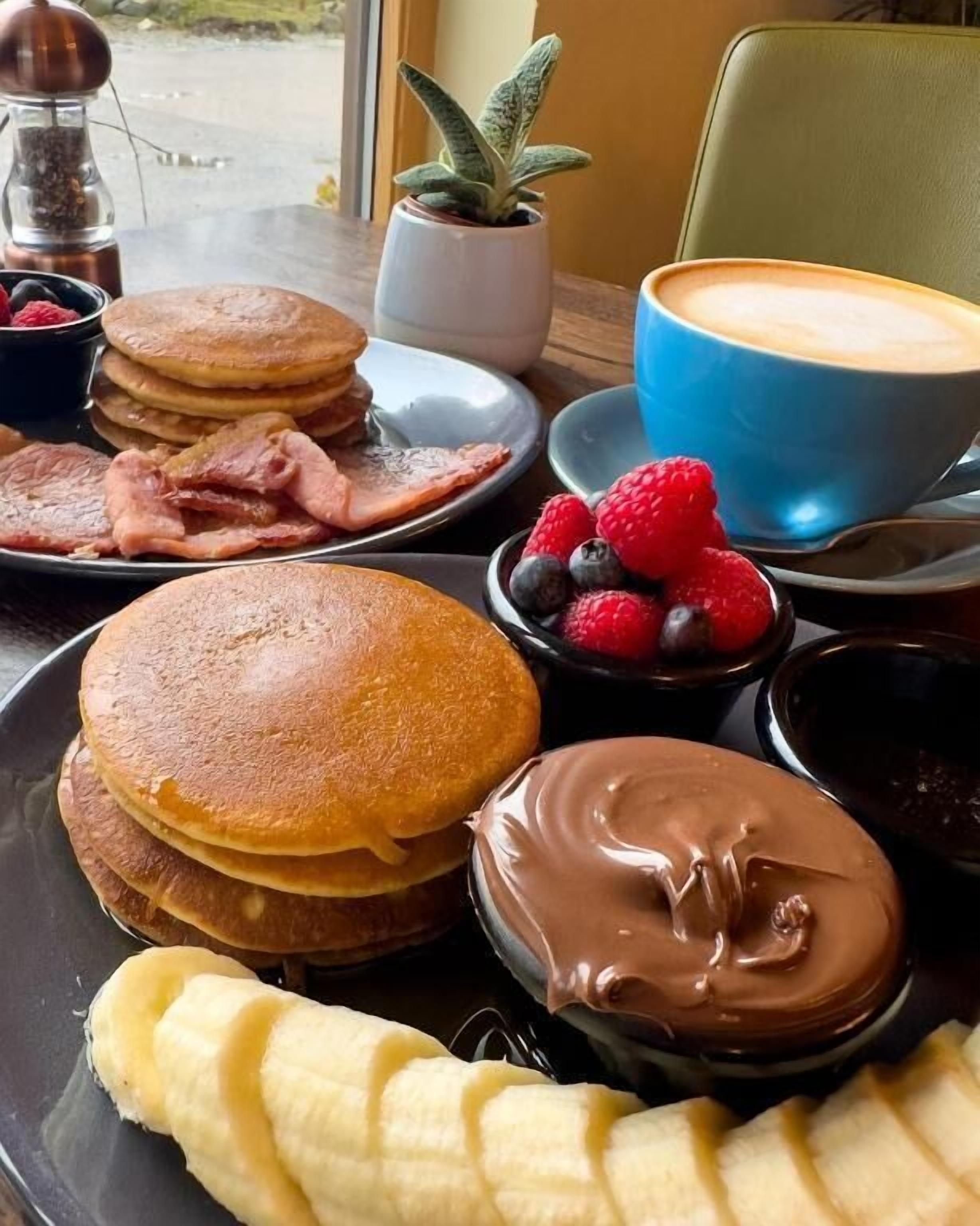 Een ontbijttafel met pannenkoeken, spek, bessen, chocolade en een koffiekop.
