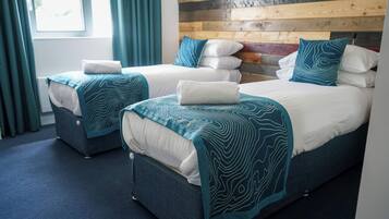 Habitación con dos camas, cada una con cubrecama azul y blanco estampado, cabecero de madera y vista al exterior a través de una ventana con cortinas.