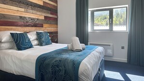 A hotel room with a wooden headboard, a large bed with white and blue bedding, a window with a view of trees, and a blue carpet. - Base Camp Hotel Nevis Range (Fort William)
