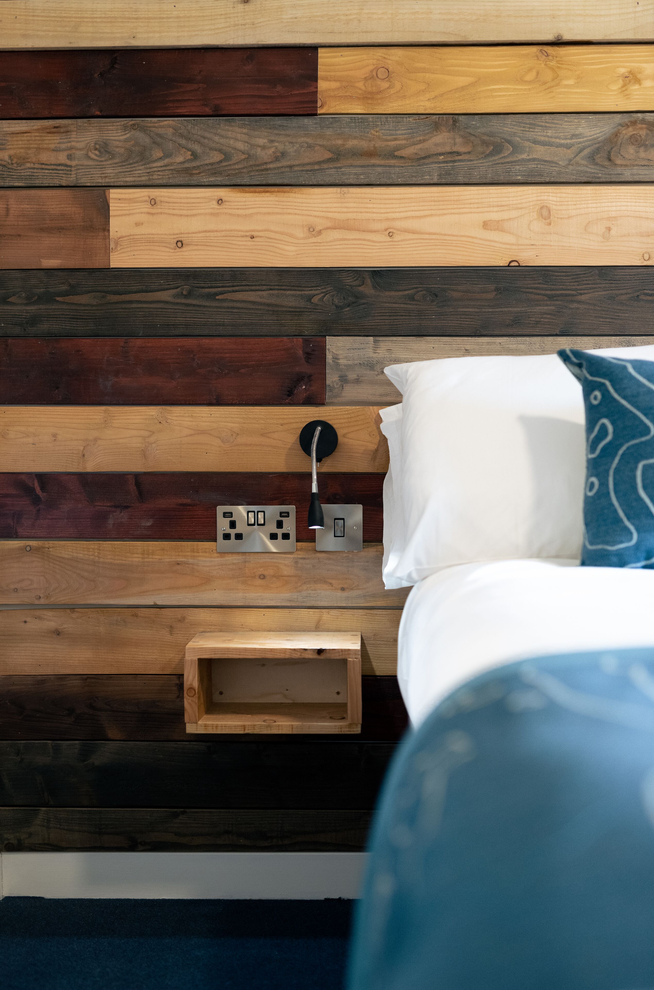Un letto con un cuscino bianco e un cuscino azzurro a motivi decorativi, un comodino con una lampada, e una parete rivestita in legno con prese elettriche.