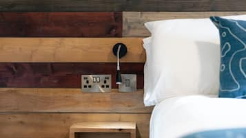 Un letto con un cuscino bianco e un cuscino azzurro a motivi decorativi, un comodino con una lampada, e una parete rivestita in legno con prese elettriche.