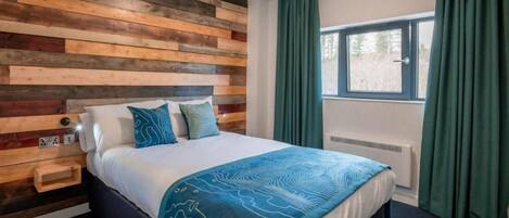 Quarto com cabeceira de madeira, cama com manta azul estampada, cortinas verdes e janela com vista para árvores.