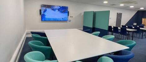 A conference room with a large table, teal chairs, a mounted TV, and a staircase in the background.