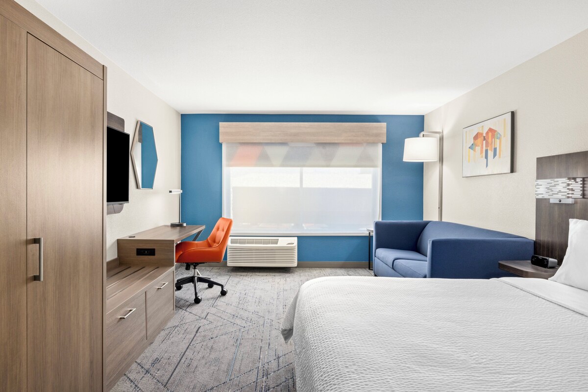 A hotel room with a bed, a desk, an orange chair, a blue sofa, and a large window with blinds.