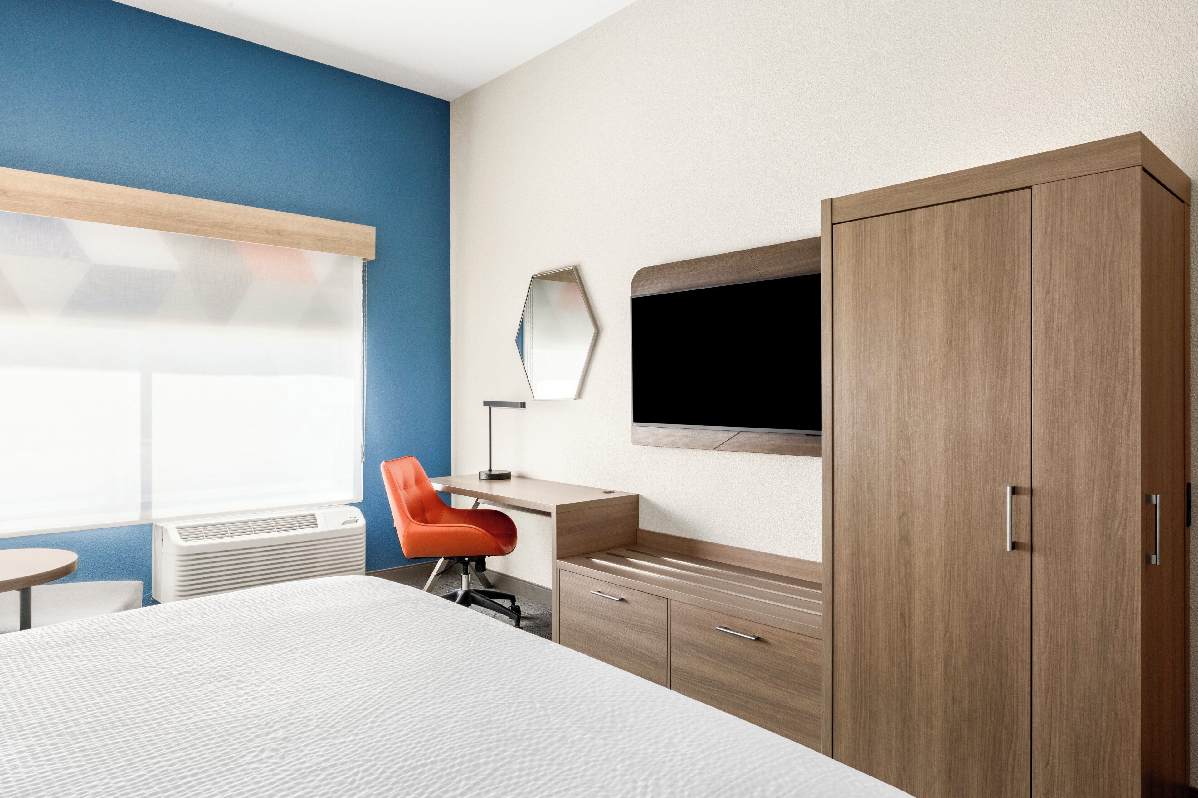 Une chambre d’hôtel avec un lit, un bureau, une chaise orange, une télévision et une armoire.
