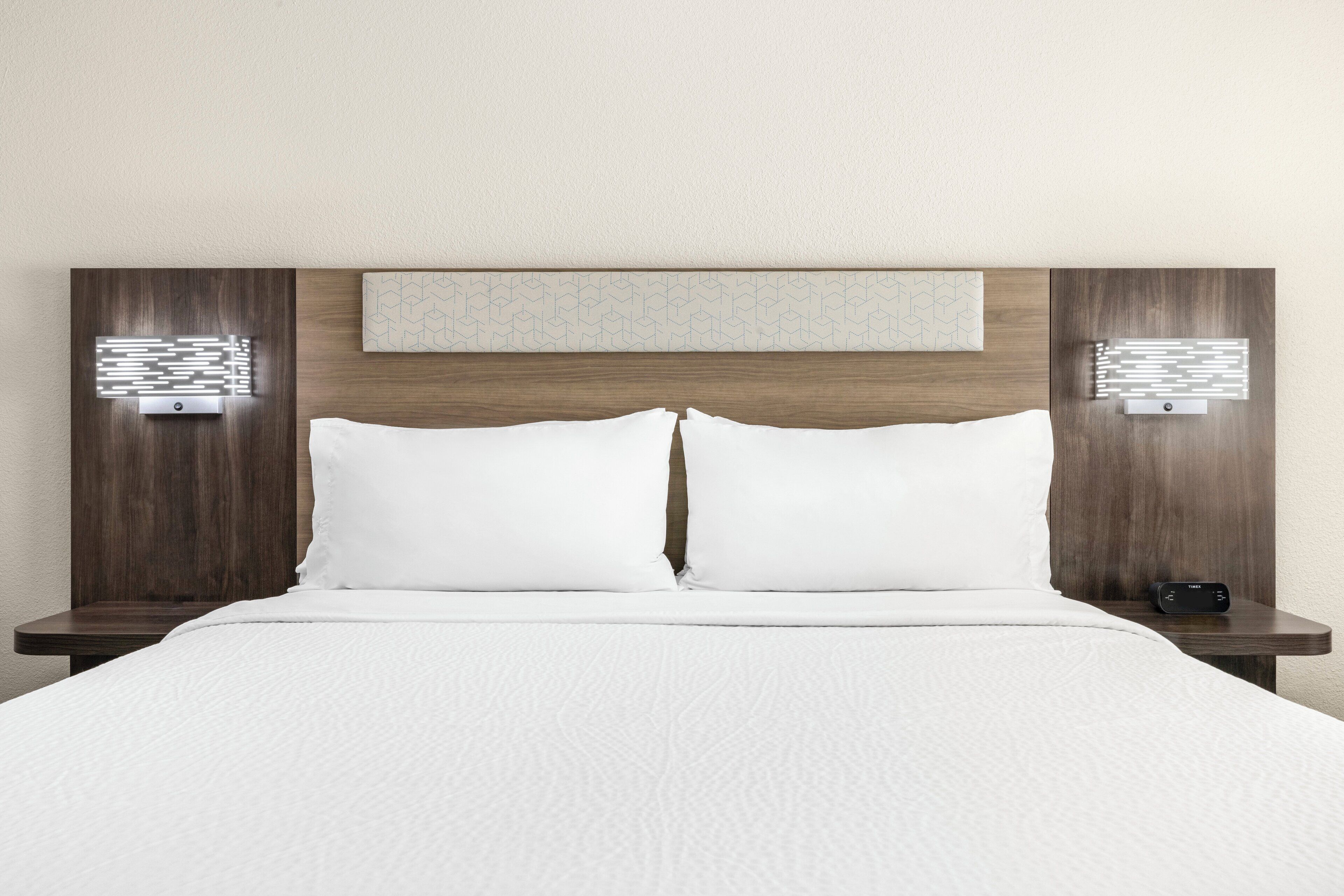 A hotel room with a wooden headboard, white bedding, and two bedside tables with wall-mounted lamps.