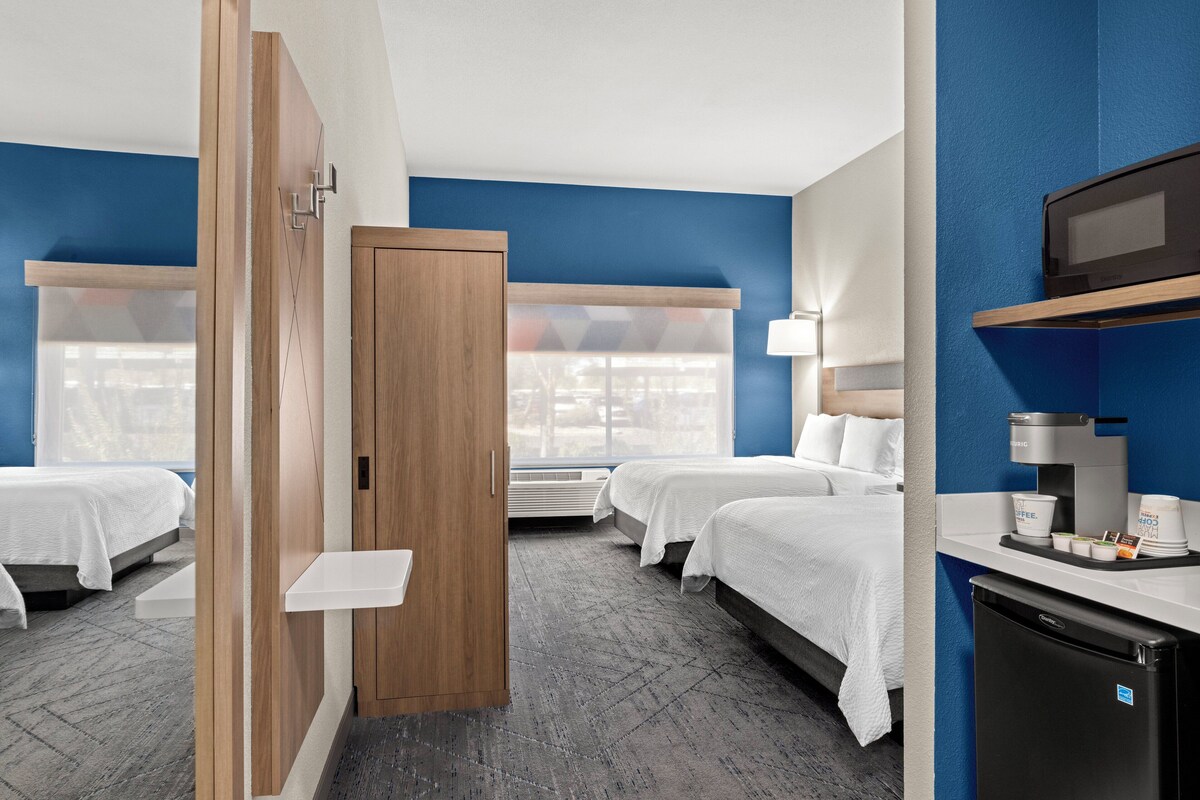 A hotel room with two beds, a wooden wardrobe, a desk with a coffee machine, and a TV.