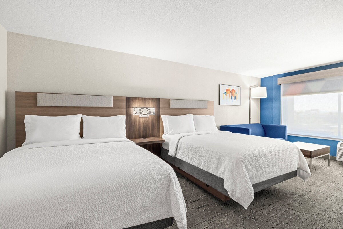 Une chambre d’hôtel avec deux lits, une tête de lit en bois, une table de chevet, une chaise bleue et une fenêtre donnant sur un paysage agréable.