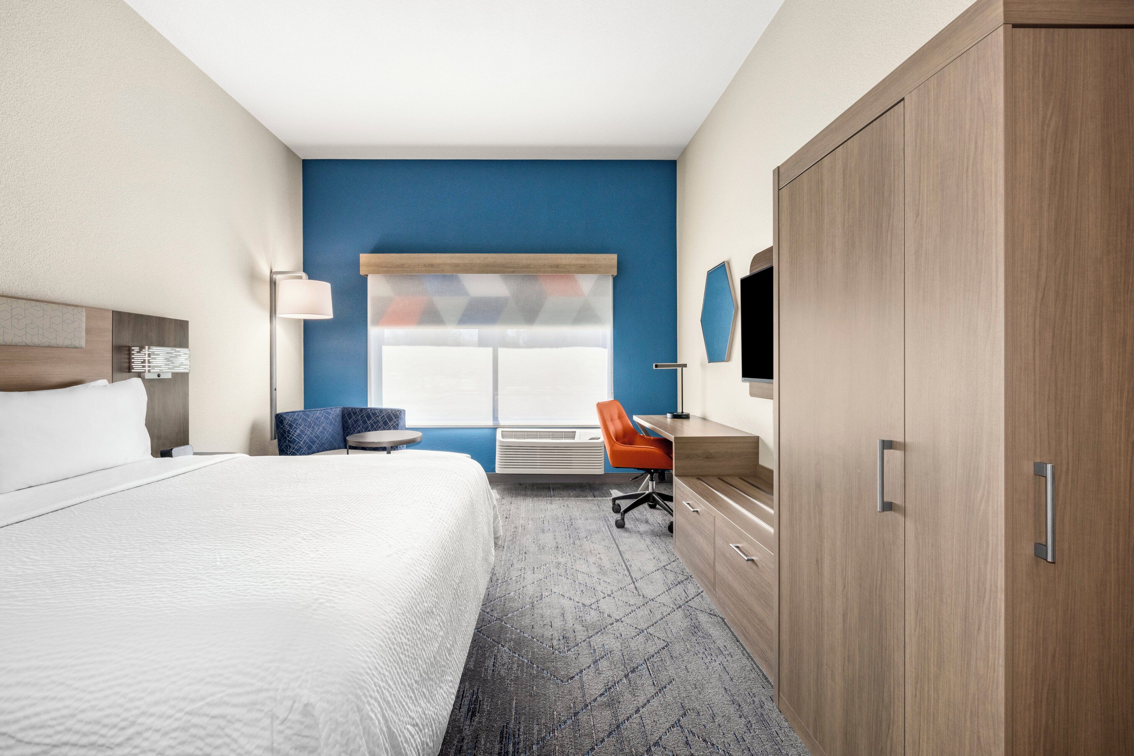 Une chambre d’hôtel avec un grand lit, un bureau, une chaise orange, une armoire et une fenêtre avec des stores.