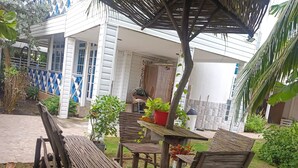 Un espace extérieur aménagé, abrité et doté de mobilier en bois et d’une table, entouré de verdure et bordé d’un bâtiment à la décoration bleue et blanche.