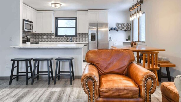 Een moderne keuken met witte kastjes, een roestvrijstalen koelkast en een ingebouwde magnetron. Er staan drie zwarte barkrukken en een bruine leren fauteuil.
