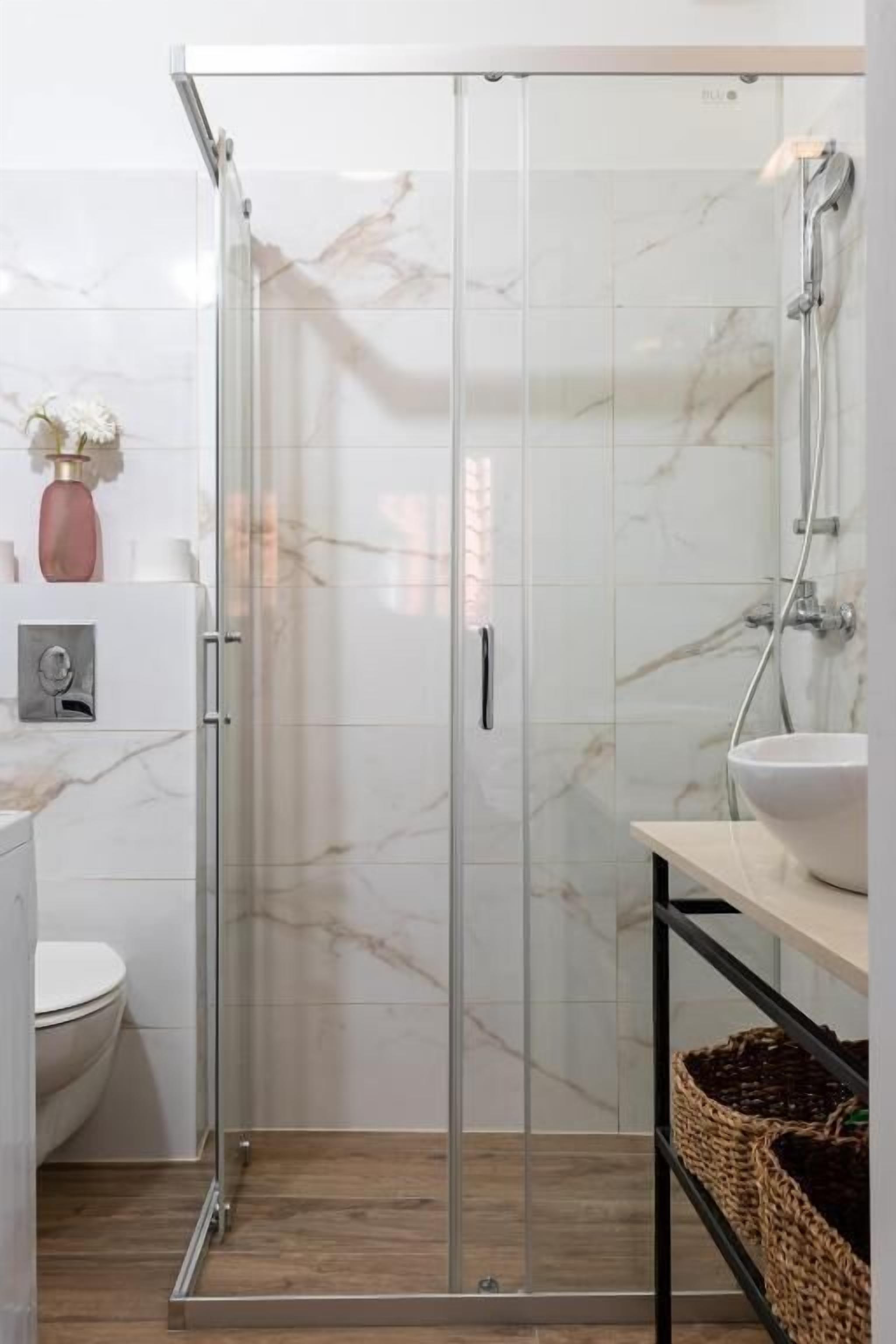 Een moderne badkamer met een douchecabine van glas, witte marmeren tegels, een houten vloer, een witte wastafel en een kleine vaas met bloemen.