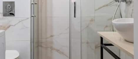 Un baño moderno con ducha de vidrio, baldosas de mármol blanco, piso de madera, lavamanos blanco y un pequeño jarrón con flores.