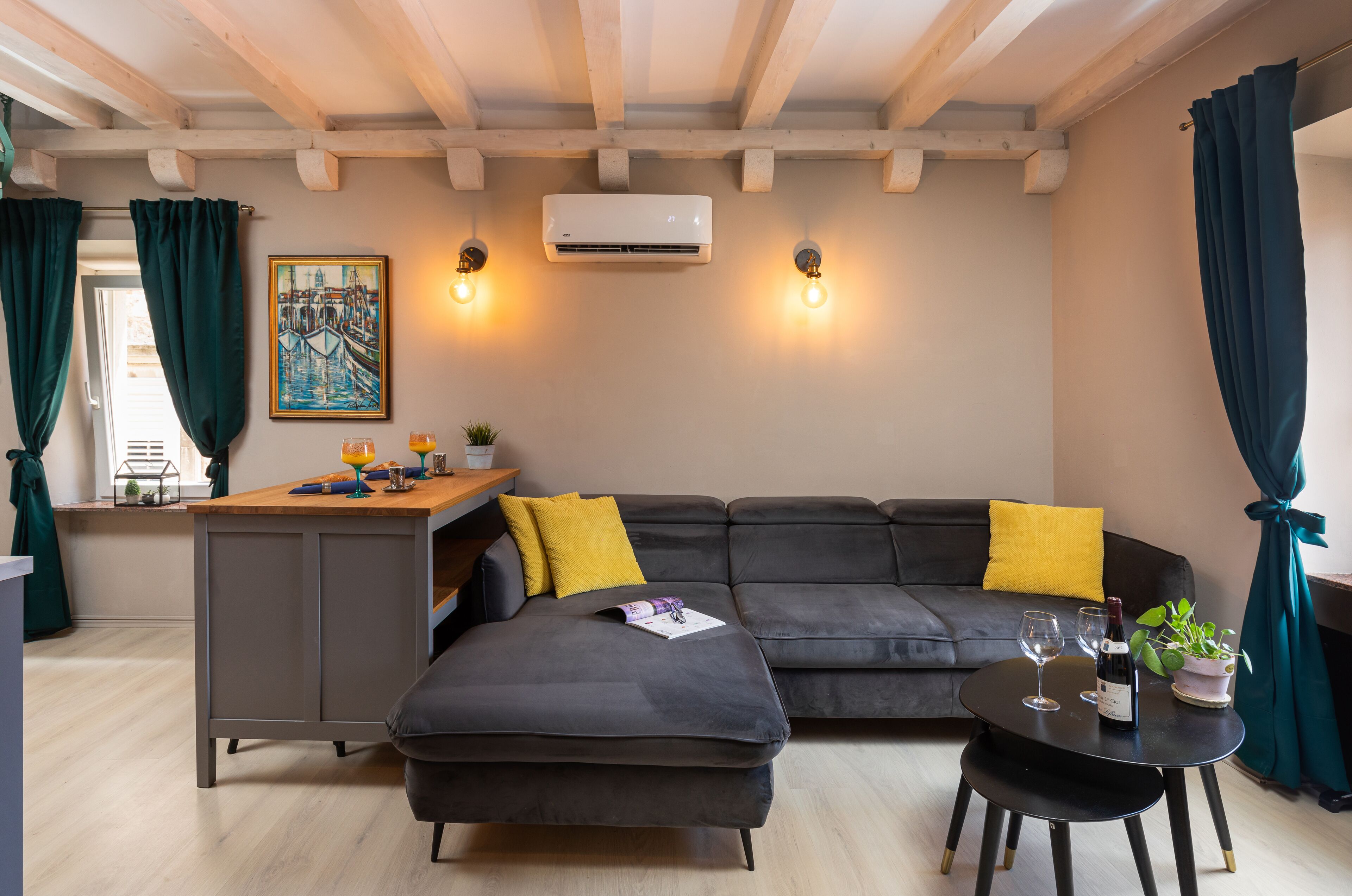 Uma sala moderna com um sofá seccional cinza escuro, uma mesa redonda pequena com duas taças de vinho e uma garrafa, uma área de cozinha com balcão e um quadro na parede.