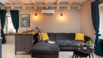 Uma sala moderna com um sofá seccional cinza escuro, uma mesa redonda pequena com duas taças de vinho e uma garrafa, uma área de cozinha com balcão e um quadro na parede.