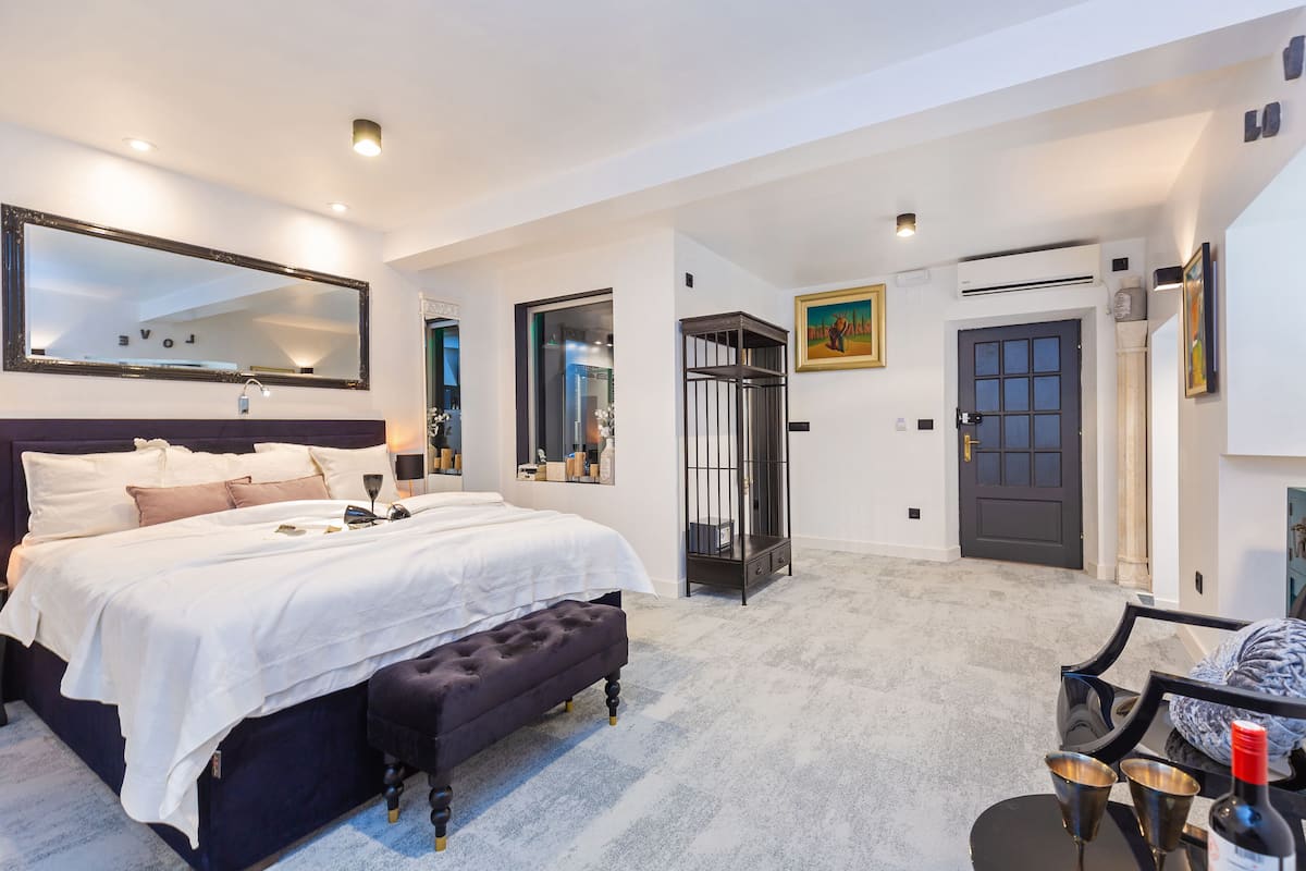 Un dormitorio amplio con una cama grande, un banco de color oscuro, un espejo y una puerta que da a otra habitación.