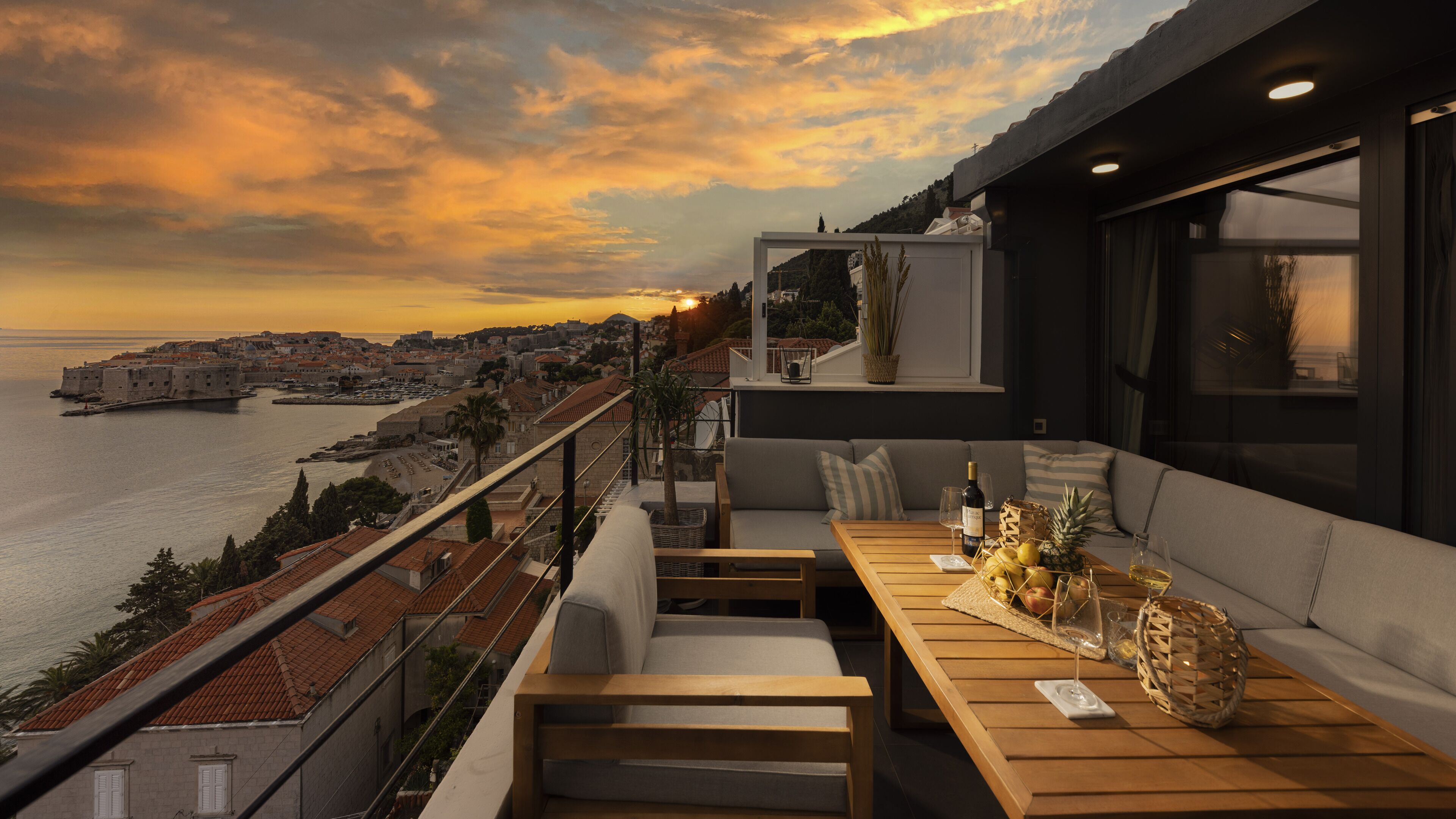 Un balcón con mesa de madera, asientos acolchados y vista a una ciudad costera al atardecer.