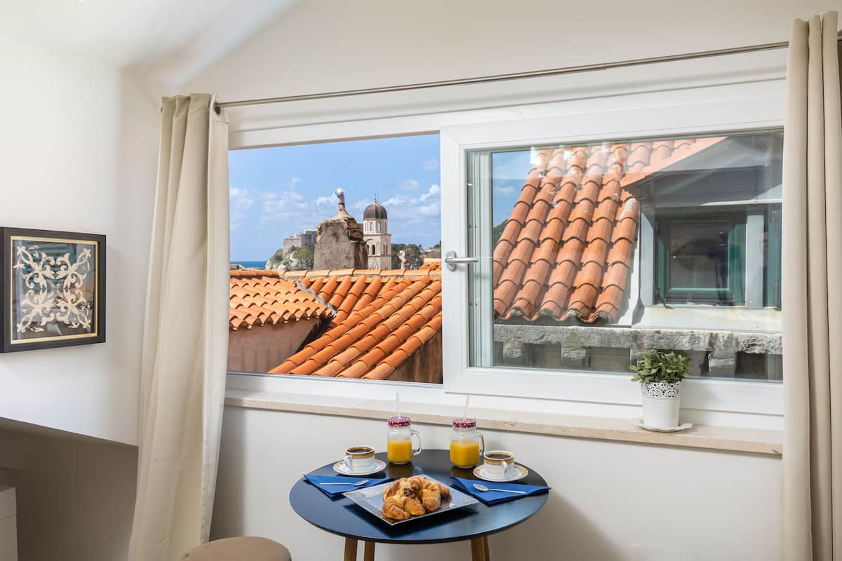 Una habitación con ventana que da a un paisaje urbano, una pequeña mesa con alimentos para el desayuno y un cuadro en la pared.