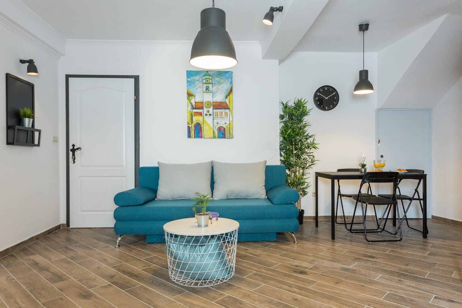 Una sala de estar con un sofá azul, una mesa redonda, una mesa de comedor con sillas, un reloj y un cuadro en la pared.