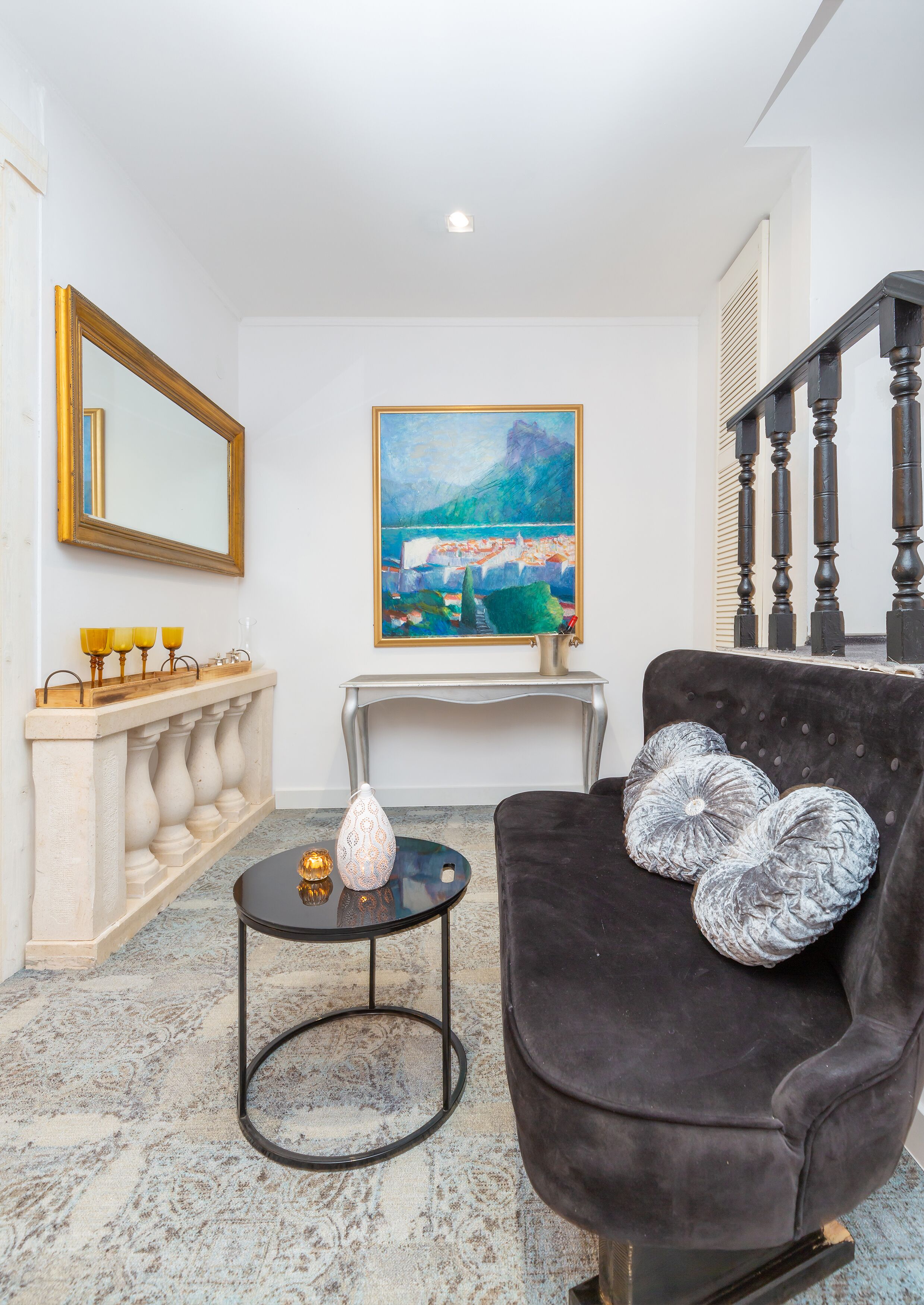 Un salon avec un fauteuil en velours sombre, une table ronde ornée d’un vase, un tableau accroché au mur et un escalier avec des rampes en bois.