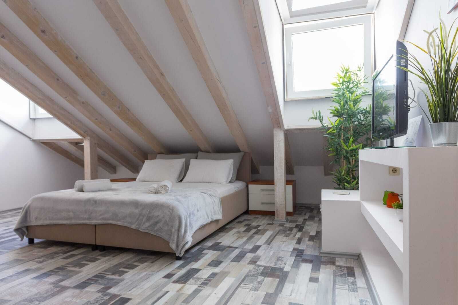 Een slaapkamer met een schuine plafond, een bed met een grijze plaid, een nachtkastje, een televisie op een plank en een potplant.