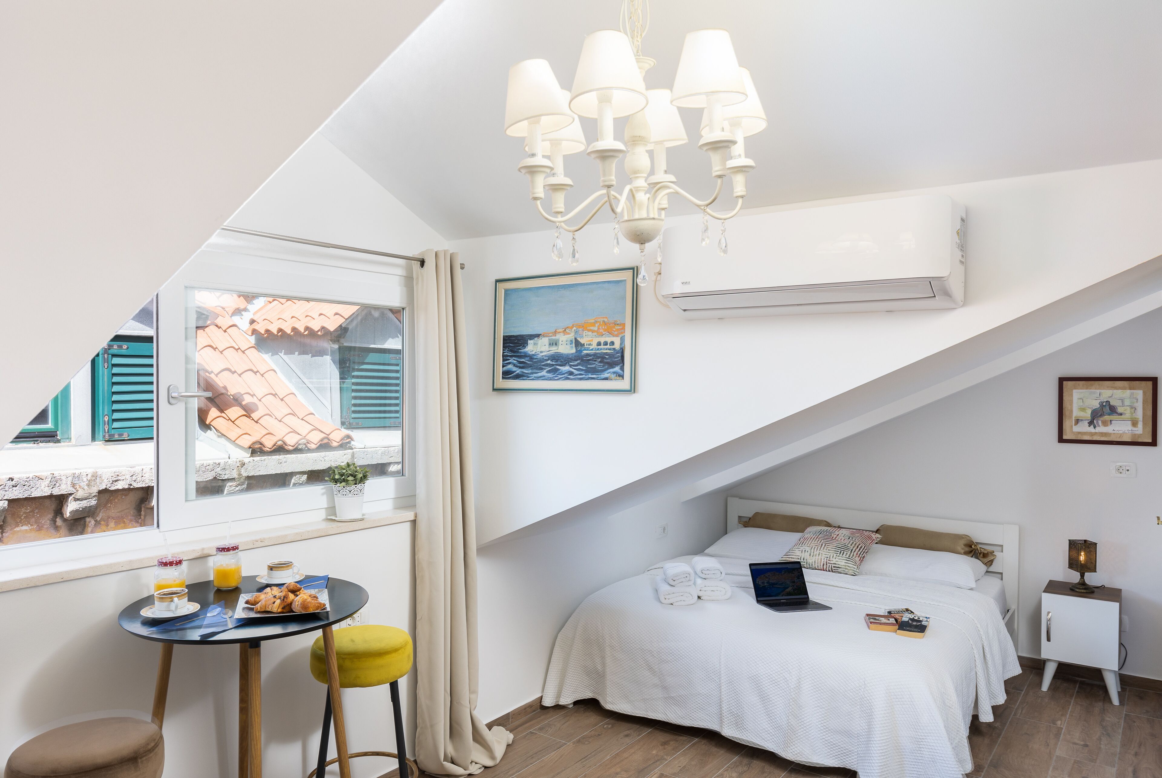 Une chambre à coucher avec un lit, une petite table sur laquelle sont disposés des éléments pour le petit-déjeuner, une fenêtre donnant sur des immeubles et un lustre.