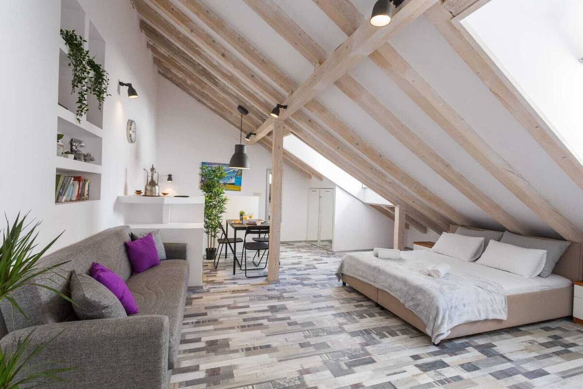 Dormitorio tipo loft con techo inclinado, cama, sofá, mesa de comedor y vista al cielo.