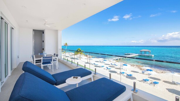A balcony with blue cushioned seating, a table, and a view of a beach with white sand, blue water, and a pier. - Rum Point Resort 206 by Grand Cayman Villas (Seven Mile Beach)