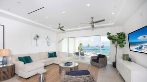 A modern living room with a white sofa, wicker chairs, a glass coffee table, and a view of the beach through large windows. - Rum Point Resort 206 by Grand Cayman Villas (Seven Mile Beach)
