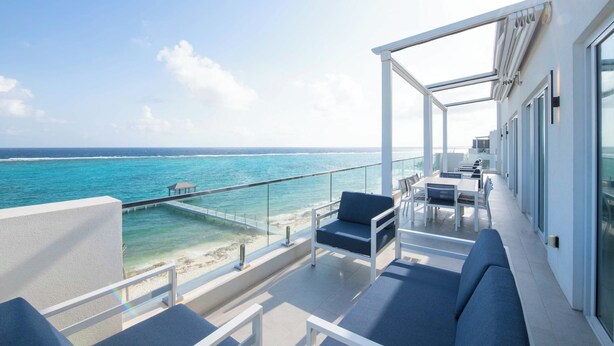 A balcony with a view of the ocean, white furniture, and a glass railing. - Rum Point Resort 401 by Grand Cayman Villas (Grand Cayman)