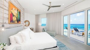 Een moderne hotelkamer met een groot bed, een plafondventilator, uitzicht op de oceaan en een balkon met lounge fauteuils.